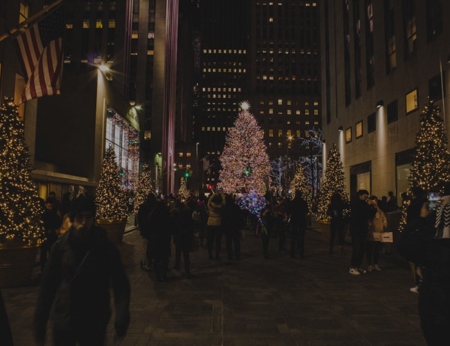 Crowds gather at night around the illuminated Rockefeller Center Christmas tree in New York City, framed by lit trees and an American flag.