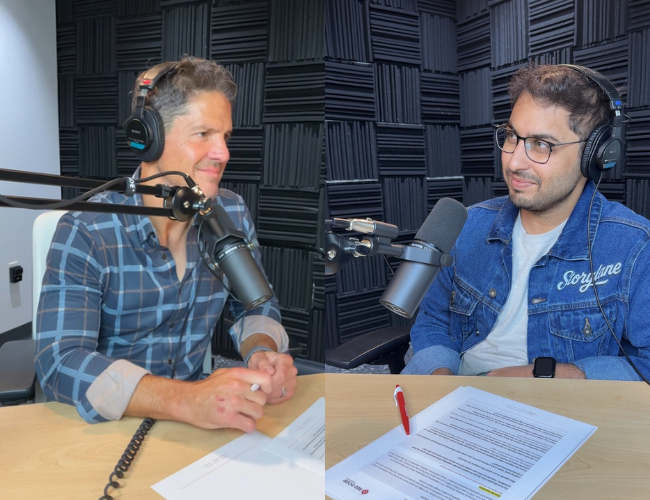 Two podcast hosts wearing headphones at microphones in a small recording booth with acoustic foam walls.