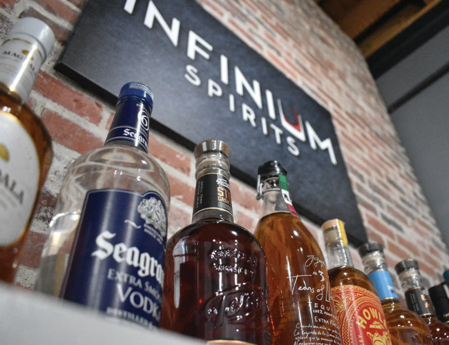 Several spirit bottles displayed on a bar shelf against an exposed brick wall with an Infinium Spirits sign.
