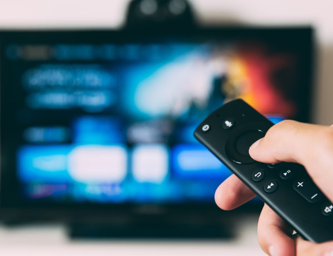 Hand holding a black streaming TV remote pointing at a blurred television screen displaying app icons.