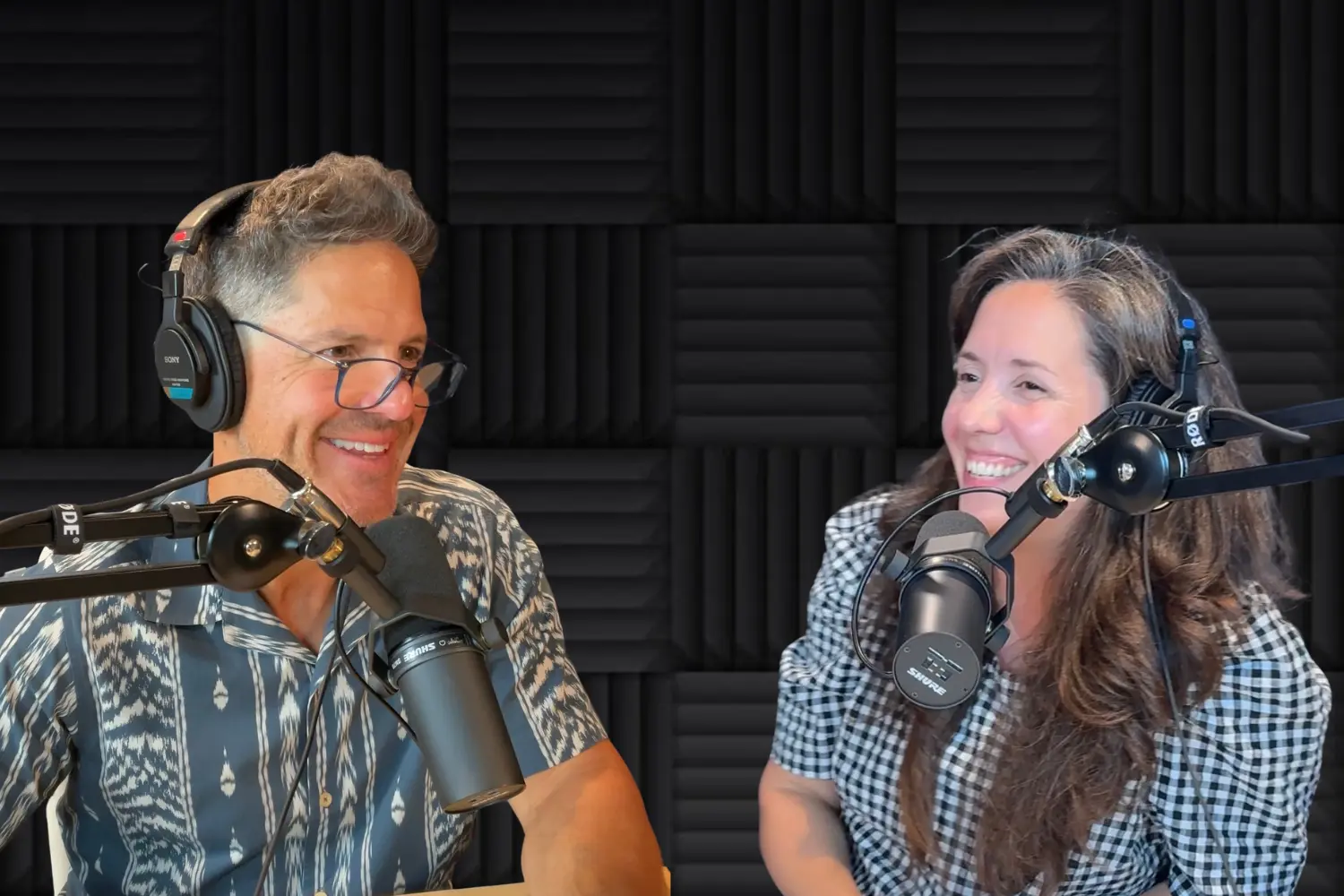 Two podcast hosts wearing headphones and smiling at microphones in a recording studio with acoustic foam walls.