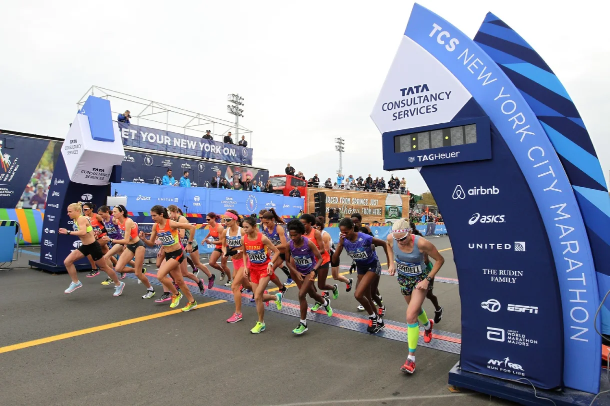 Women runners surge from the start line at the TCS New York City Marathon, with sponsor banners for ASICS, Airbnb, and United visible on the arch and surrounding signage.