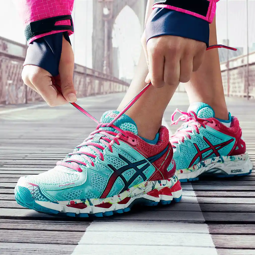 Close-up of a runner tying the laces on a colorful ASICS running shoe in teal, pink, and red, with the Brooklyn Bridge visible in the background.