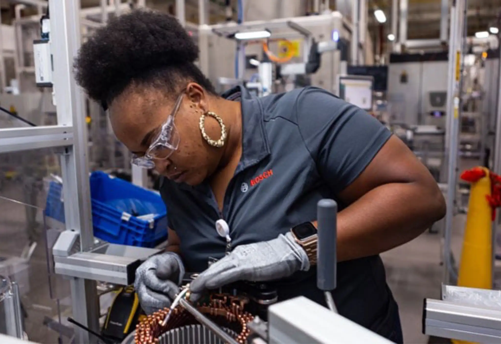 A Bosch-uniformed technician wearing safety glasses and gloves carefully works on an electrical component on an assembly line at a manufacturing facility.