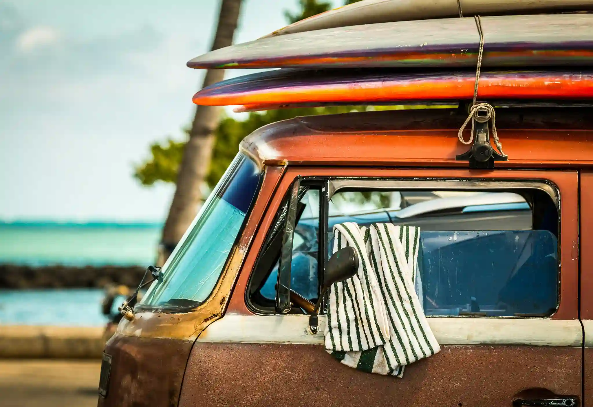Close-up of the cab of a weathered vintage van with colorful surfboards strapped to the roof, parked near a tropical beach with palm trees in the background.