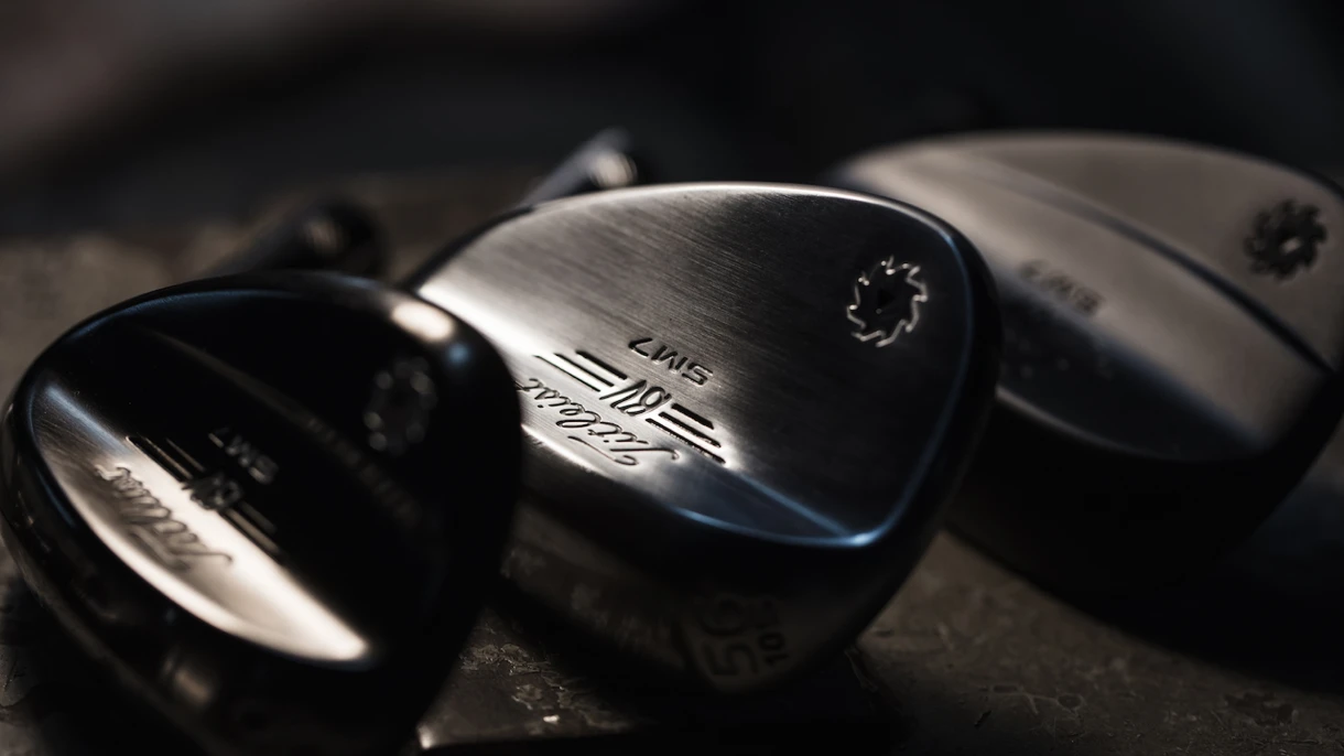 Close-up of three Titleist Vokey SM7 wedge heads resting on a dark surface, with dramatic studio lighting highlighting the brushed metal finish and engraved Titleist and Vokey branding.