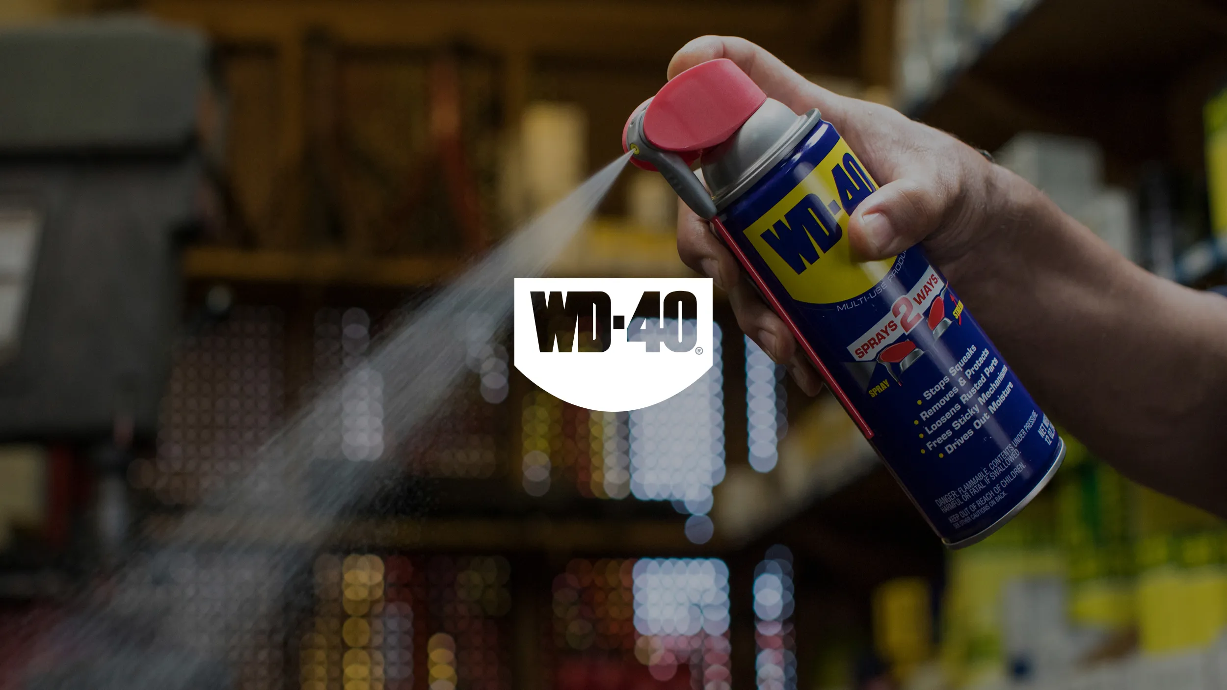 A hand sprays WD-40 from a blue and yellow can with a red nozzle. A fine mist is visible, conveying action. Shelves blur in the background.