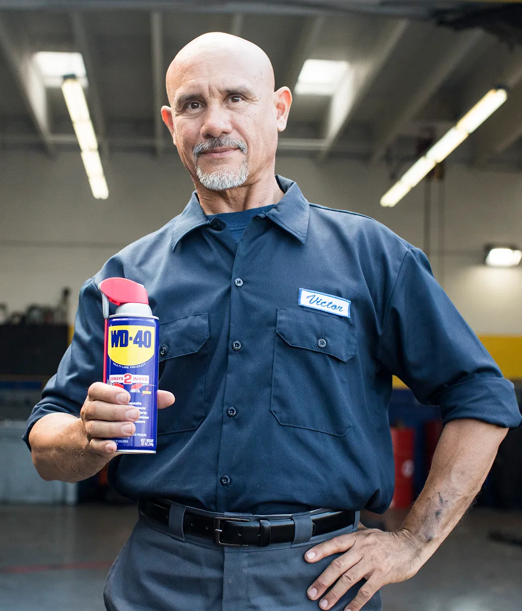 A mechanic in a blue uniform stands confidently in a garage, holding a can of WD-40. The setting is industrial, with bright overhead lights.