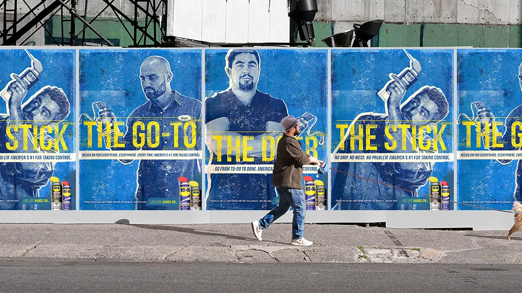 A series of three blue-toned posters on a street wall feature men holding WD-40 cans with text like "The Stick" and "The Go-To." A person walks by.