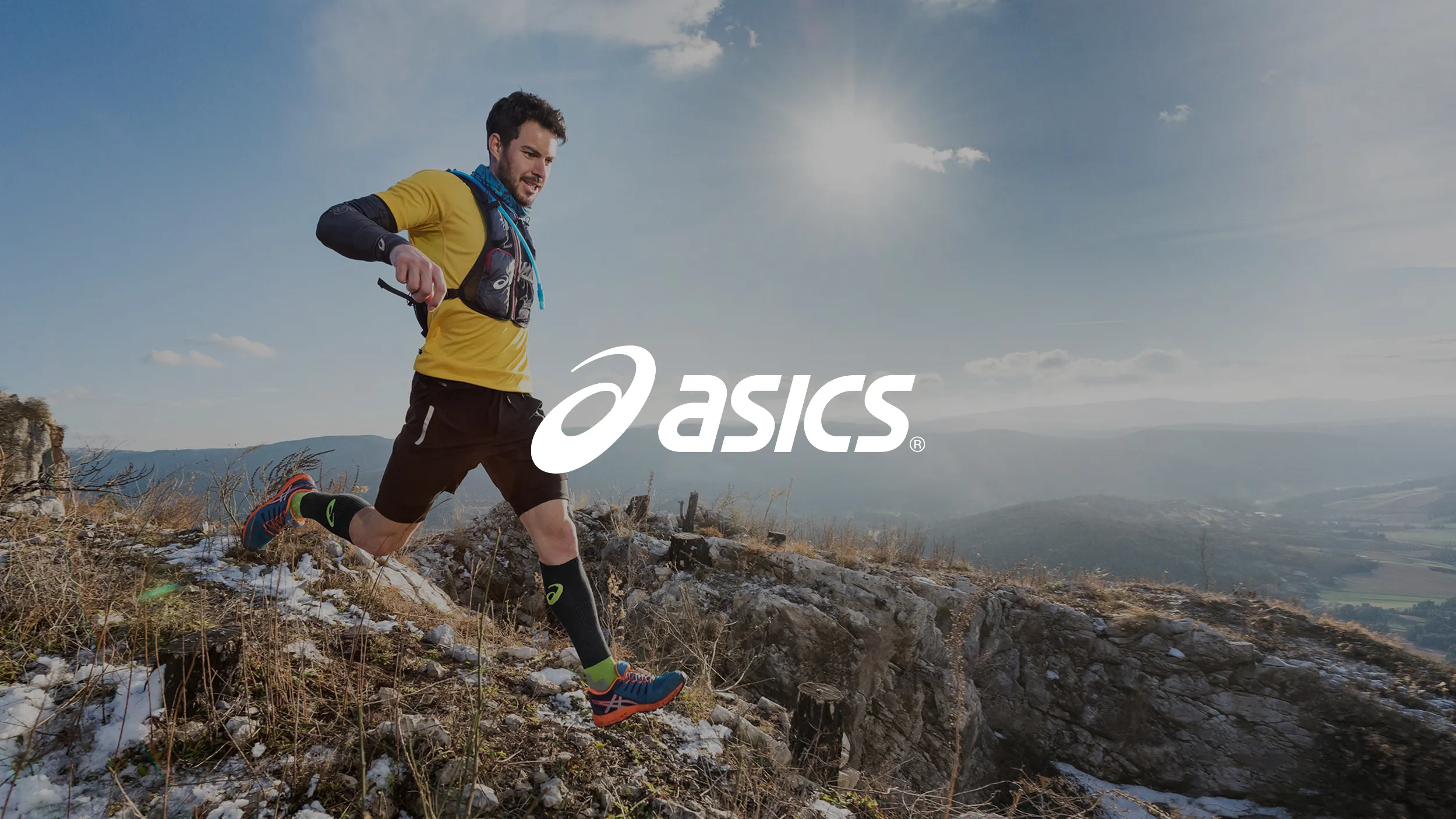A man in athletic gear runs along a rocky, snow-dusted trail, exuding energy and determination. The sun shines brightly in the clear sky above, with "Asics" logo centered.