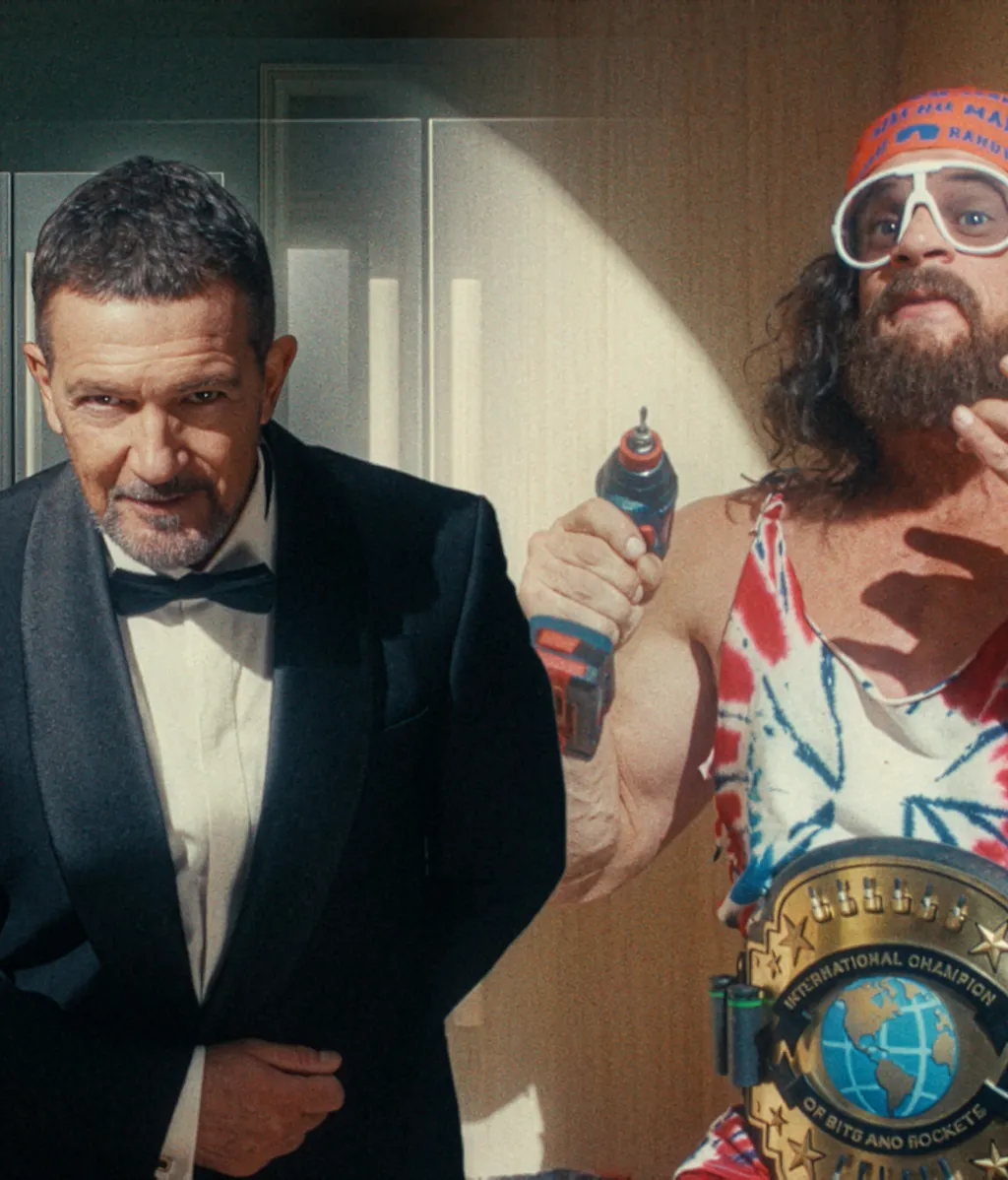 A man in a tuxedo and a flamboyant character in a tie-dye tank top, headband, and oversized sunglasses holding a Bosch power drill and wearing a wrestling-style championship belt, posed together in a humorous Bosch ad still.