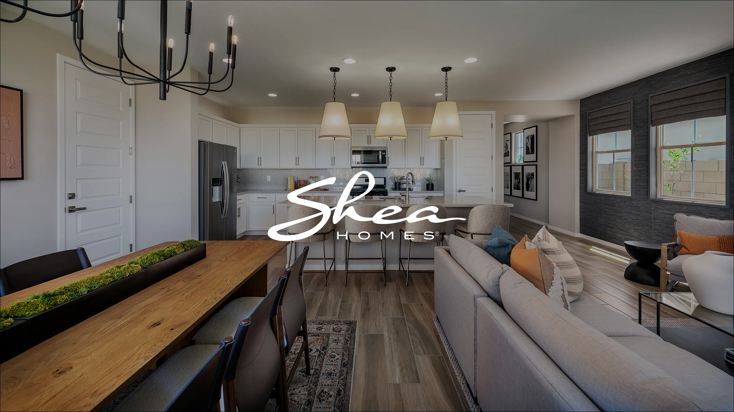 Modern open-concept living area with a dining table and kitchen. Features stylish decor, recessed lighting, and neutral tones. Shea Homes logo overlayed in the center of the image.
