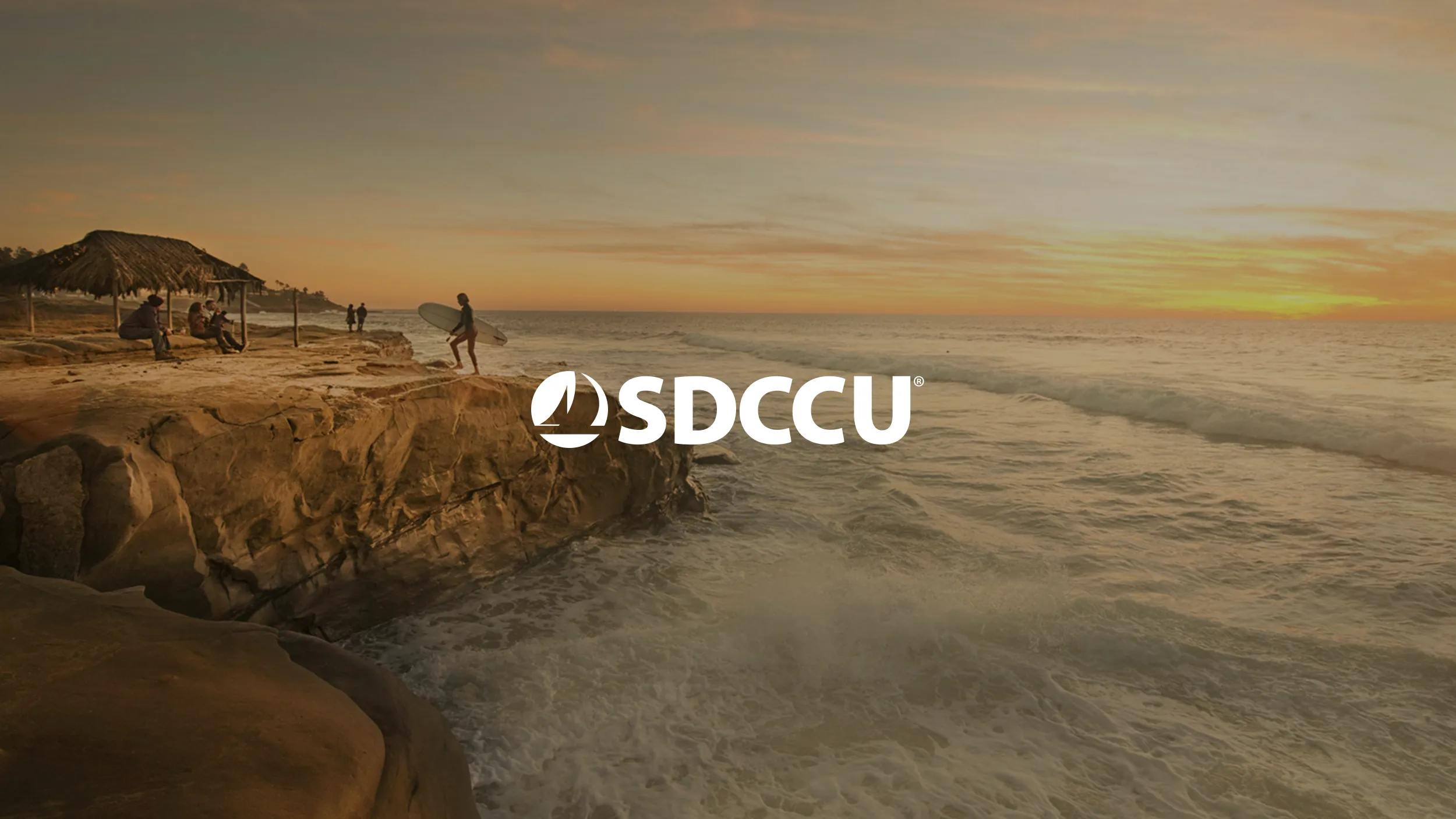 A warm golden sunset over the rocky cliffs of Sunset Cliffs in San Diego, with a surfer carrying a surfboard and a small group gathered beneath a thatched palapa shelter. Ocean waves crash below the sandstone bluffs. The white SDCCU logo is centered over the scene.