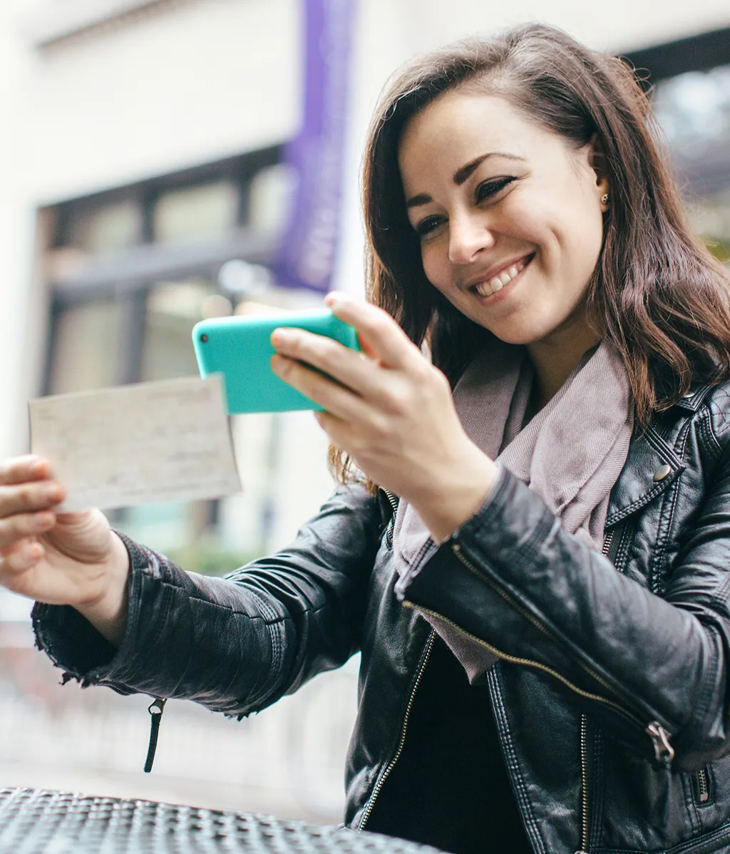 A smiling woman with dark hair wearing a black leather jacket uses a teal smartphone to photograph a check for ▎ mobile deposit while seated at an outdoor café patio.