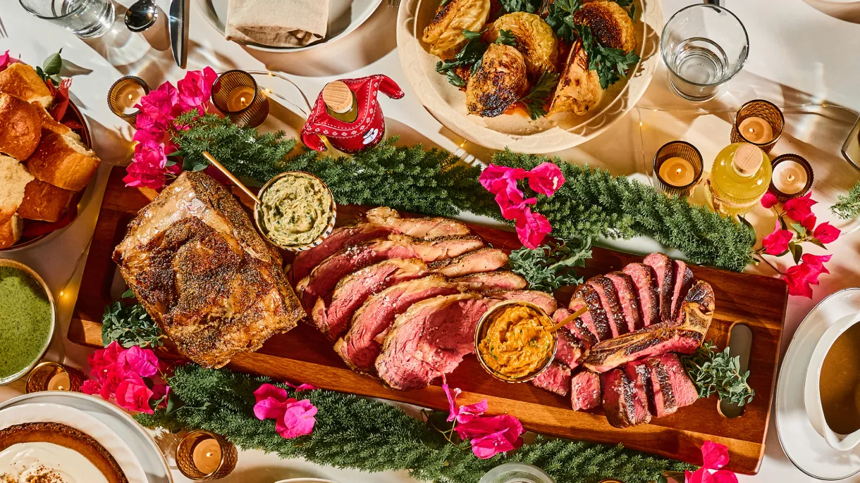 A wooden platter with sliced roast beef, flanked by sauces and fresh herbs, surrounded by vibrant pink flowers and various side dishes, creating a festive atmosphere.
