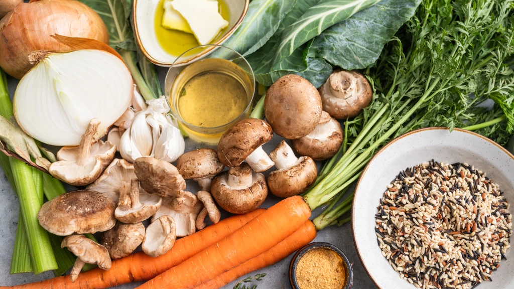 A vibrant assortment of vegetables and ingredients on a table, including mushrooms, carrots, onions, garlic, rice, butter, and oil, conveying freshness.