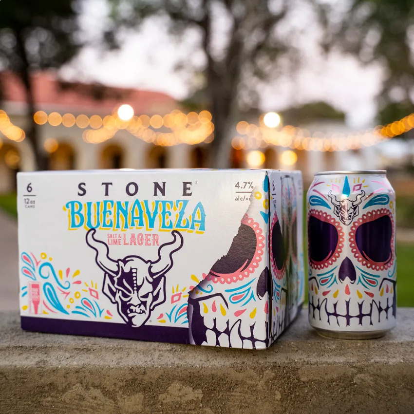 Stone Buenaveza Salt & Lime Lager 6-pack and can with Day of the Dead skull design on a stone ledge with string lights in the background.