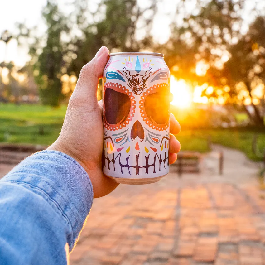 A hand holding a Stone Buenaveza Salt & Lime Lager can against a golden sunset in an outdoor park setting.
