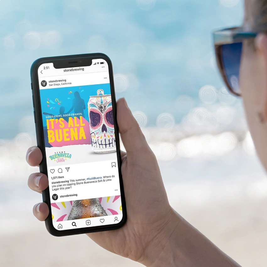 A person at the beach holding a smartphone displaying the Stone Brewing Instagram page with a Buenaveza "It's All Buena" post.