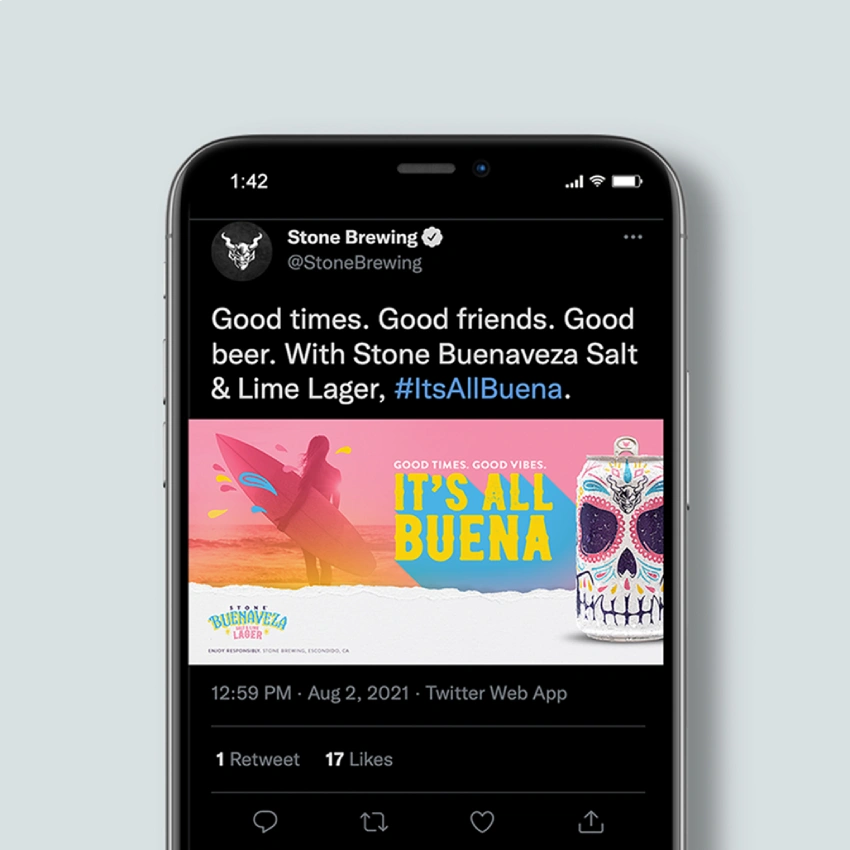 A smartphone displaying a Stone Brewing tweet promoting Buenaveza Salt & Lime Lager with an "It's All Buena" surfer ad image.