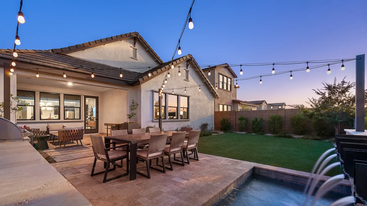 Backyard patio at twilight with a wooden dining set, string lights above, a grassy lawn, and a small pool with fountains. Warm and inviting ambiance.
