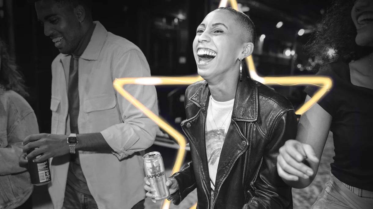 A black-and-white image shows three friends laughing and holding canned Sapporo drinks on a city street at night. A glowing yellow star overlays the center.