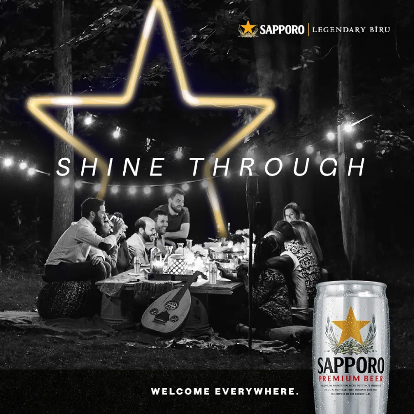 A group of people enjoy a picnic in a forest at night, with string lights and a glowing star above. Text reads "Shine Through." Sapporo beer can in the foreground.