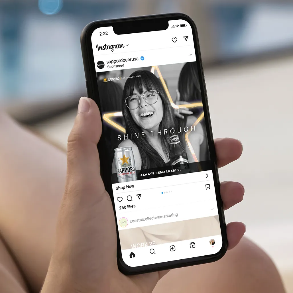 Close-up of a hand holding a phone, displaying an Instagram post. The post shows a smiling woman with glasses, a Sapporo beer, and the text "Shine Through."