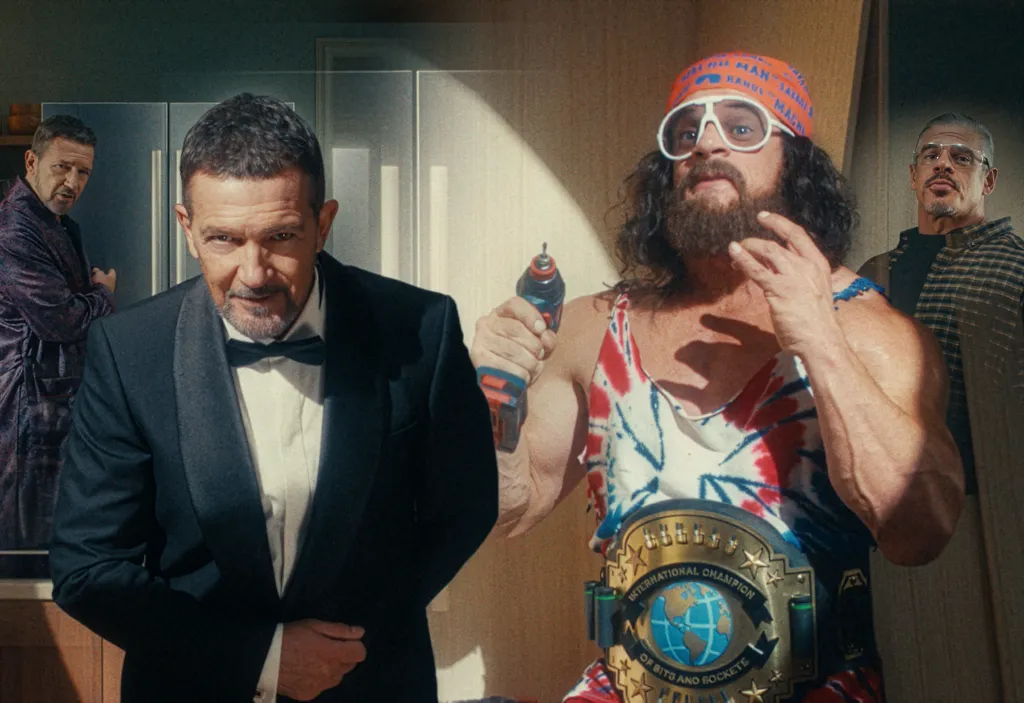 A man in a tuxedo stands confidently, flanked by a wrestler with a drill and two casually dressed men in the background.