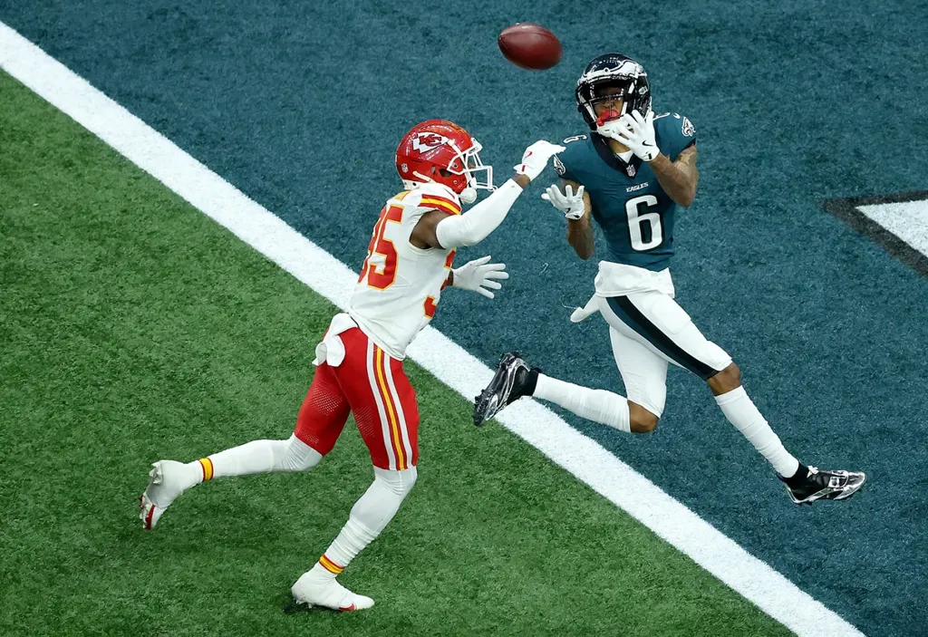 Eagles player in green leaps to catch football in end zone, pursued by Chiefs defender in red during intense game moment.
