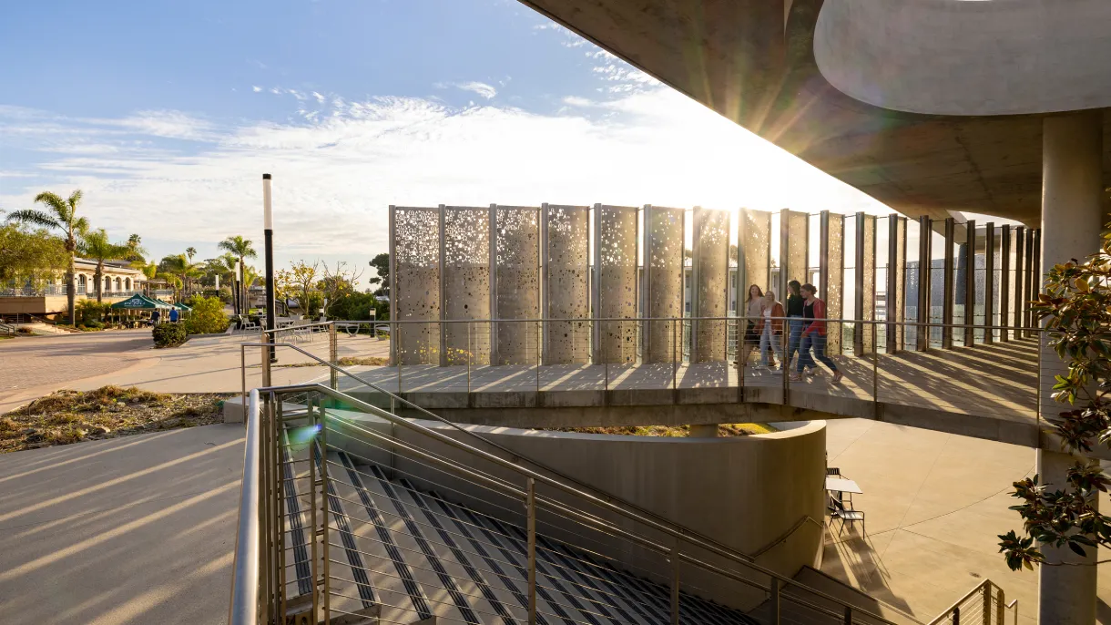 People walk on a sunlit pathway at Point Loma Nazarene University beside artistic panels, with palm trees and a clear sky in the background.