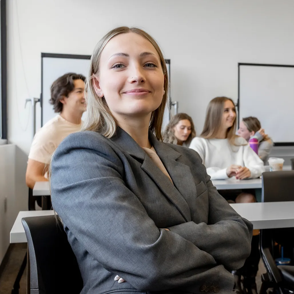 Confident woman in a gray blazer stands in front of a classroom, with students seated and engaged in the background.
