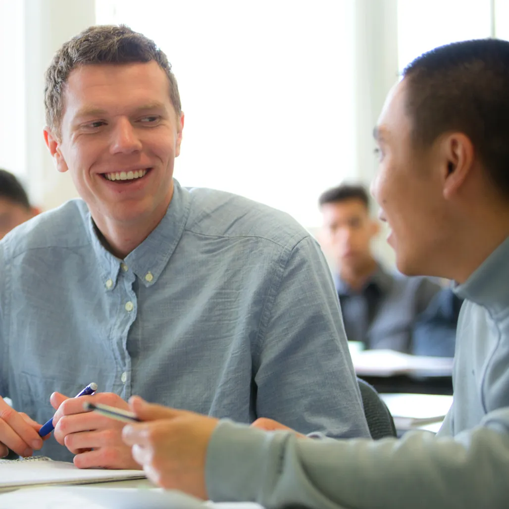 Two students in a classroom share a friendly conversation, smiling and holding pens, with others blurred in the background.