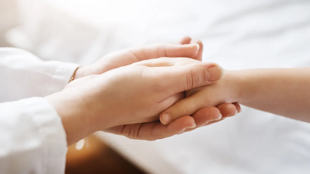 Two hands gently holding another hand, conveying warmth and support. The background is softly blurred, emphasizing the compassionate gesture.