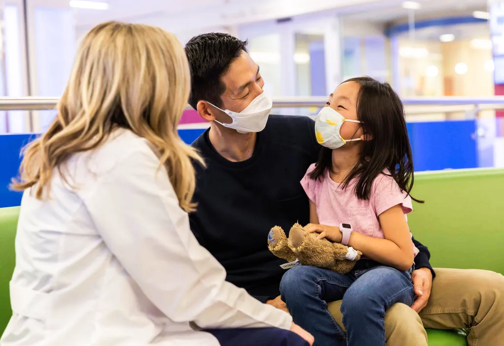 A woman in a white coat converses with a masked man and child. The child holds a teddy bear, conveying a warm, reassuring atmosphere in a hospital.