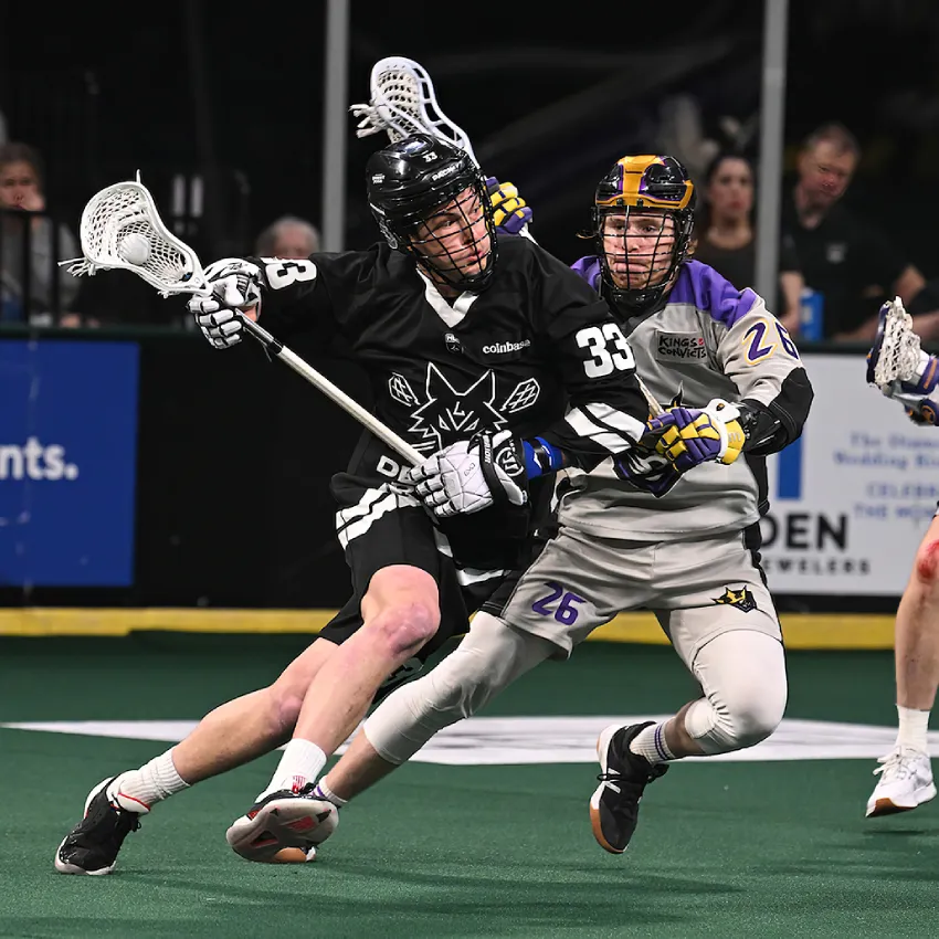 Desert Dogs player #33 in black jersey cradles the ball while shielding the stick from a defender from the Kings in purple and gray.