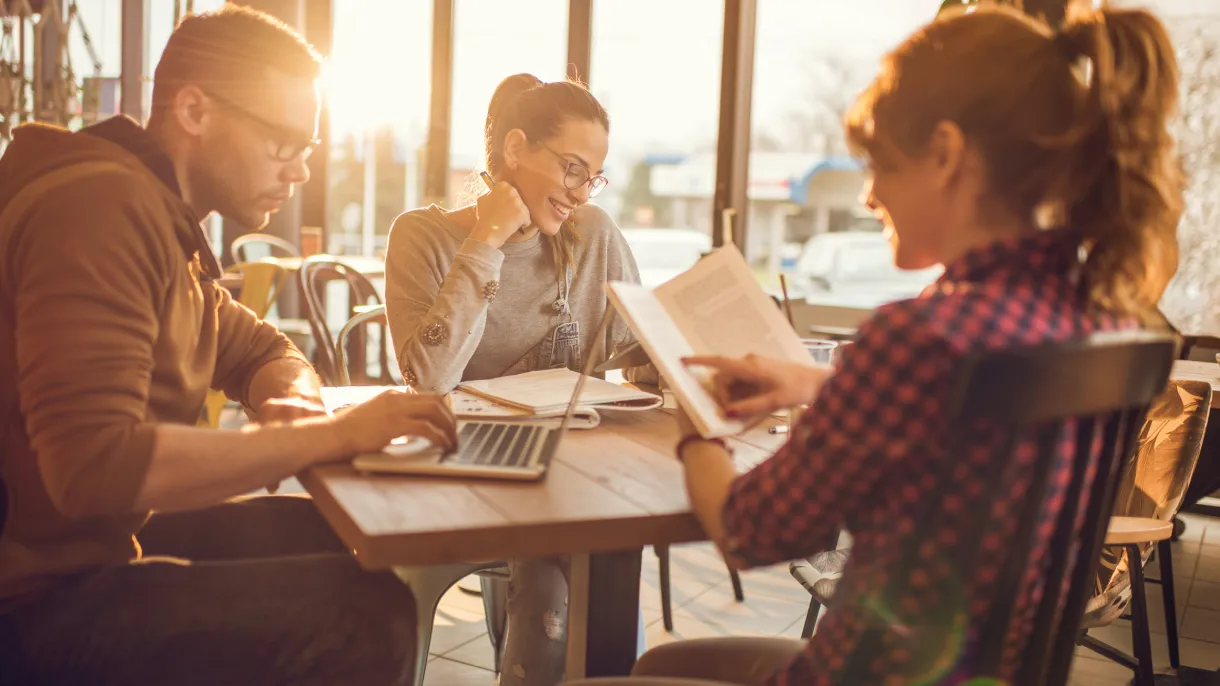Three students studying with a laptop, books and notes at a sunlit cafe table, smiling and collaborating in warm golden window light.