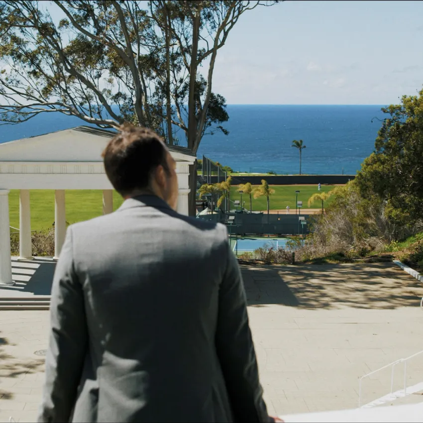 Man in a gray suit, seen from behind, looking out from PLNU's campus toward the rotunda, athletic fields and the Pacific Ocean horizon.