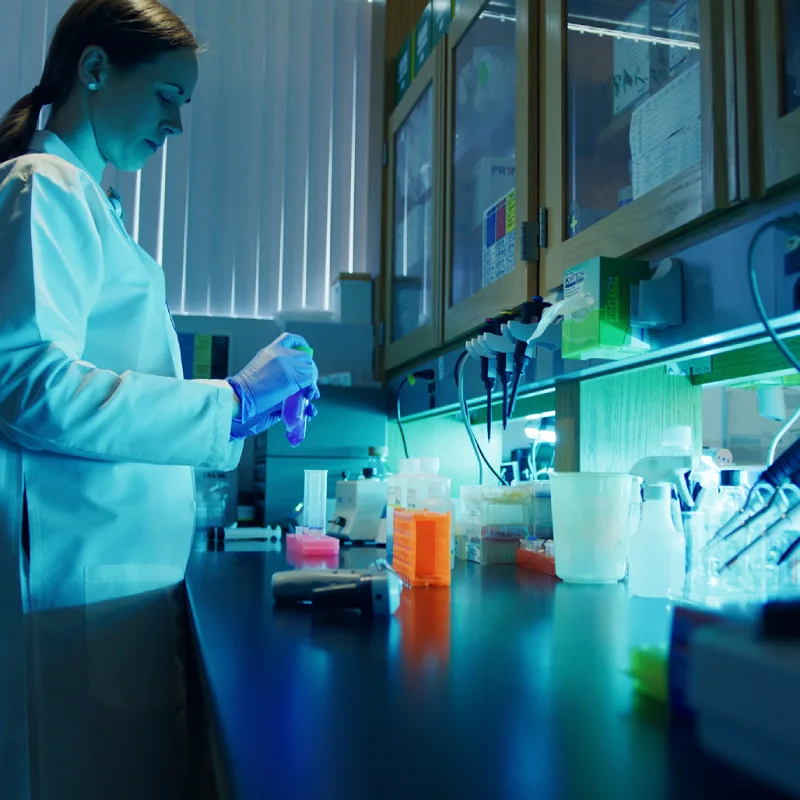  Female researcher in white coat and blue gloves works at a teal-lit lab bench with reagents and pipettes.