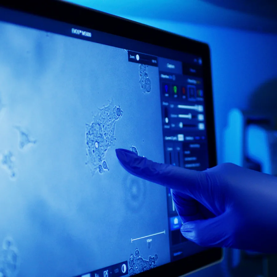Gloved finger points at cell imagery on an EVOS M5000 microscope screen bathed in blue light.