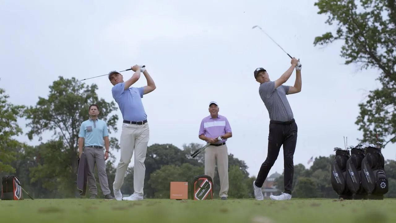 Two professional golfers mid-swing on a practice tee while a coach and another player watch, with Titleist golf bags lined up nearby. 