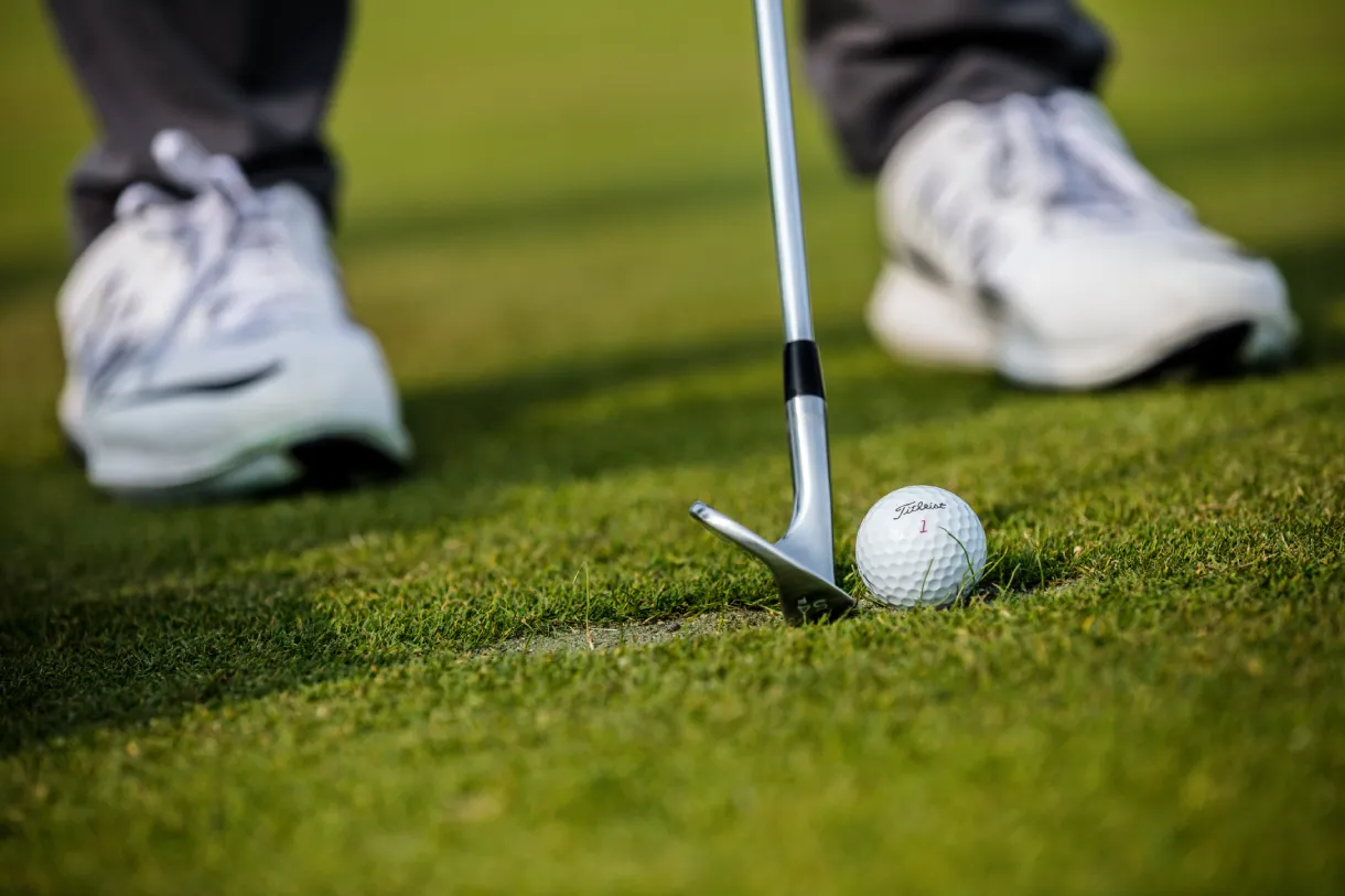 Close-up of a Titleist golf ball on grass beside a wedge club head, with a golfer's white shoes blurred in the background.