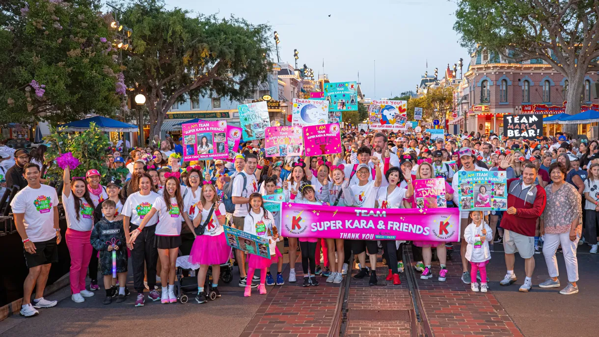 A large, joyful group gathers on a theme park street, holding colorful signs and a banners. The backdrop includes trees and classic park buildings, and the atmosphere is festive and lively.