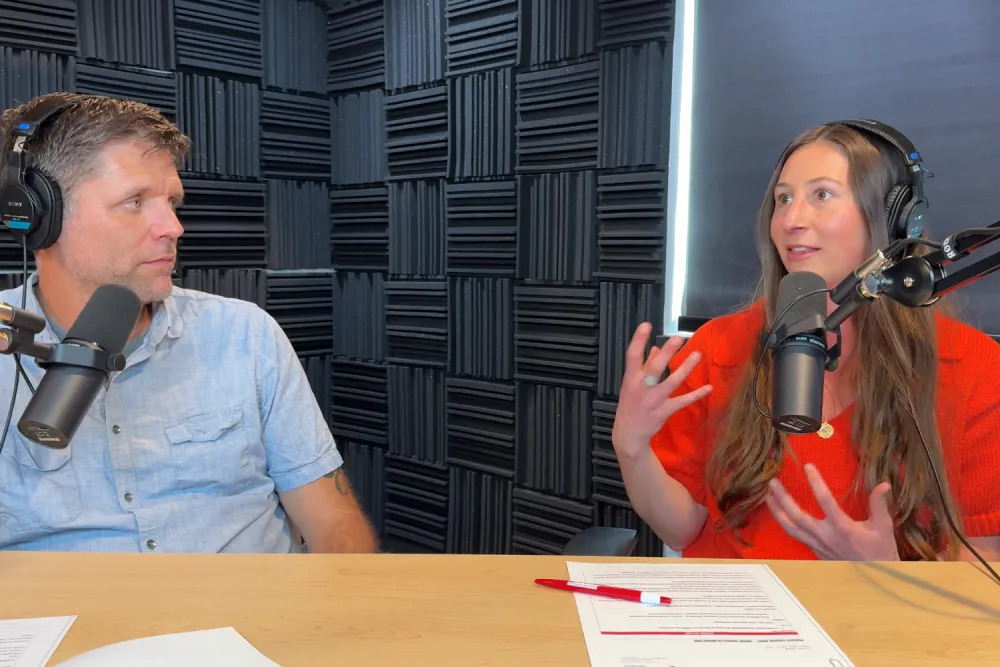 Two podcast hosts in headphones record at a studio table with mics and acoustic foam walls; the woman in red gestures while speaking as the man in blue listens.
