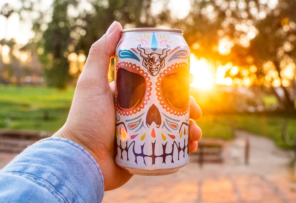 Hand holding a Stone Buenaveza Salt & Lime Lager sugar-skull can in a park at sunset, with golden light flaring through the trees.