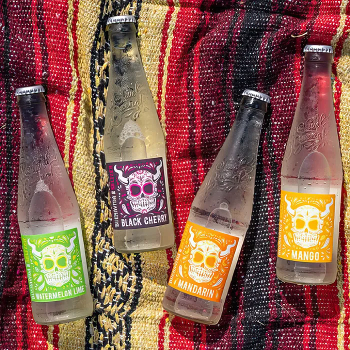 Four Stone Buenaveza bottles with green, pink, orange and yellow sugar-skull labels arranged on a striped Mexican blanket.