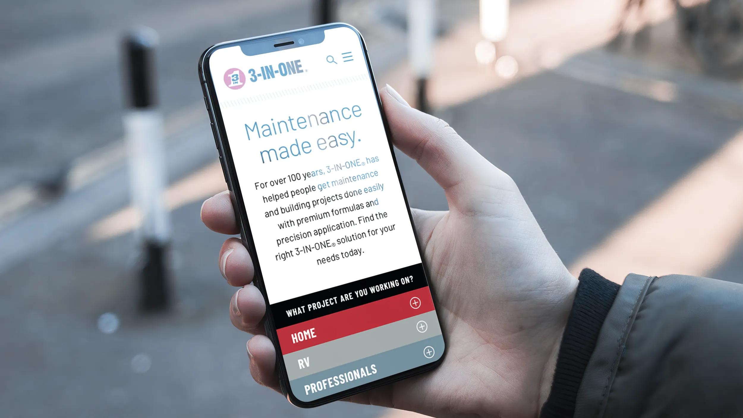 Hand holding a smartphone displaying the 3-IN-ONE mobile website with "Maintenance made easy" headline and Home, RV, and Professionals project categories.