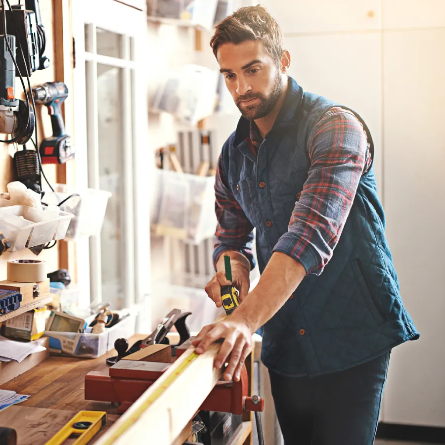 Bearded man in a flannel shirt and vest measuring a wooden board with a tape measure at a sunlit home workshop bench.