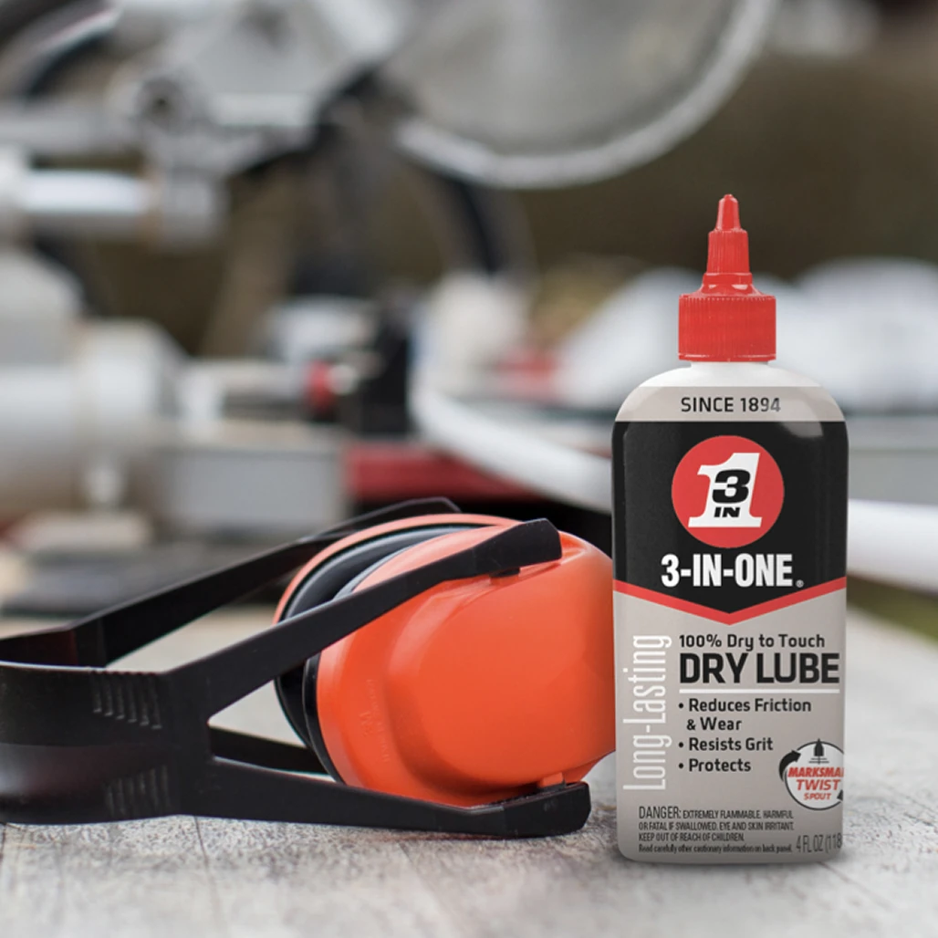 Bottle of 3-IN-ONE Long-Lasting Dry Lube next to orange safety earmuffs on a workbench with a miter saw blurred behind.
