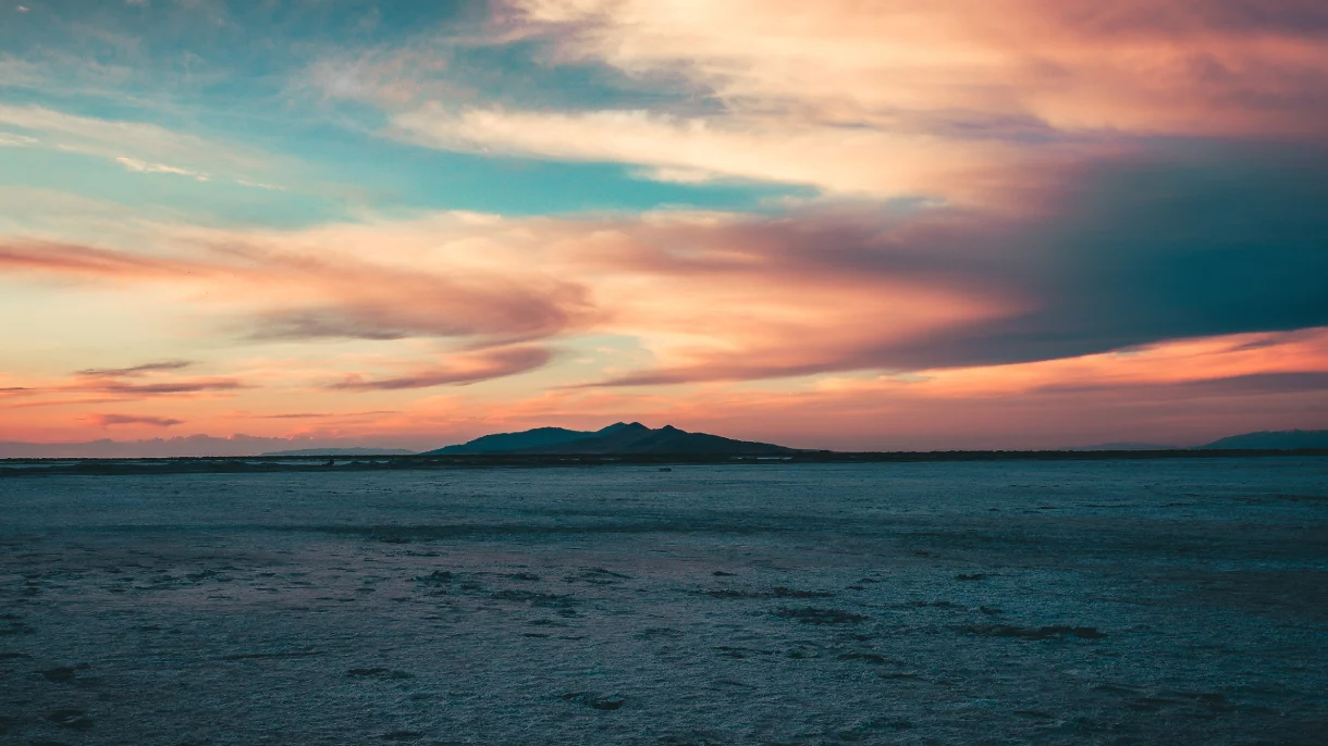 Pastel sunset sky over a quiet coastal landscape with a distant mountain silhouette across the water. 